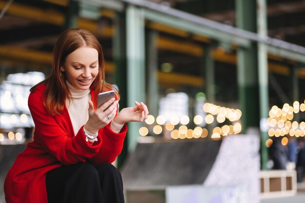 La giovane donna europea soddisfatta in cappotto rosso tiene il telefono cellulare legge la notifica connessa a Internet wireless ha tempo libero posa all'aperto attende che qualcuno si diverta con la comunicazione online