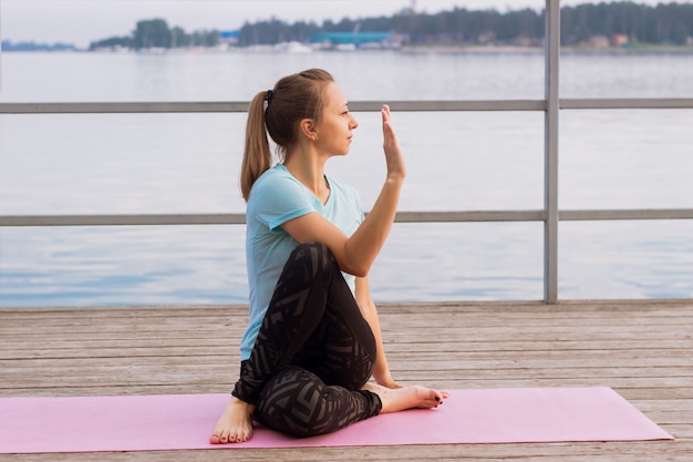 La giovane donna fa yoga su una stuoia rosa su un molo in riva al mare