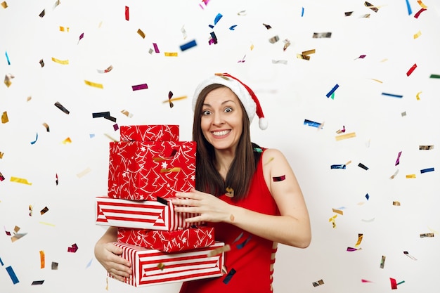 La giovane donna felice abbastanza caucasica si è vestita in vestito rosso e cappello di Natale che tiene scatole regalo su sfondo bianco e coriandoli colorati. Ragazza della Santa con il presente isolato. Concetto di vacanza di Capodanno 2018.