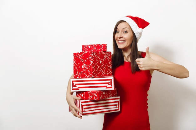 La giovane donna felice caucasica sorridente si è vestita in vestito rosso e cappello di Natale che tiene i contenitori di regalo e che mostra pollice su su fondo bianco. Ragazza della Santa con il presente isolato. Concetto di vacanza di Capodanno 2018.