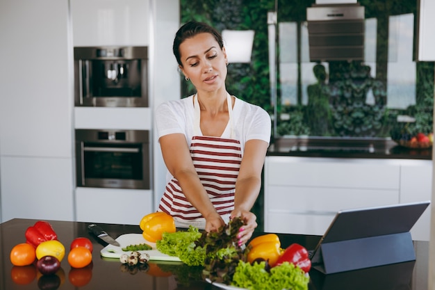 La giovane donna felice che tiene le verdure in mano in cucina con il computer portatile sul tavolo