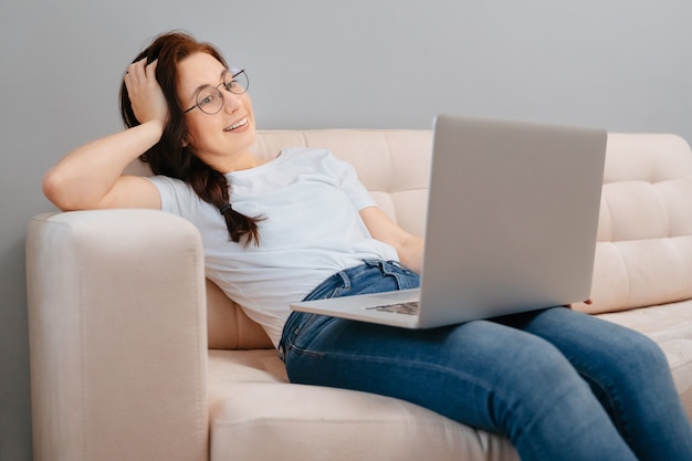 La giovane donna felice con gli occhiali sta riposando a casa sul divano ordinando merci online su un laptop con...