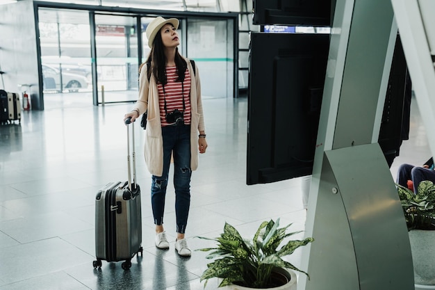 La giovane donna felice è arrivata all'aeroporto per partire e iniziare il viaggio. ragazza che cammina in piedi al cartello orario orario del volo con i bagagli. signora in abbigliamento casual guardando le informazioni del monitor all'interno.