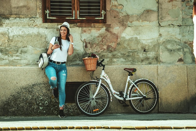 La giovane donna felice e pensierosa si sta godendo una giornata di sole estivo, appoggiandosi a un muro di una vecchia casa, accanto alla bici con un cesto di fiori.