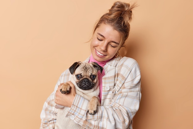 La giovane donna felice e premurosa porta il cane carlino tiene la sua zampa abbraccia con amore sorride felicemente indossa abiti casual isolati su sfondo beige. Persone animali domestici e concetto di relazione