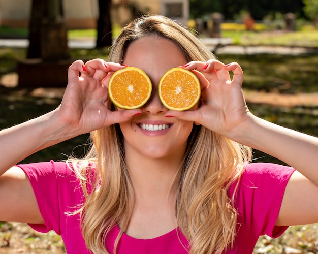 La giovane donna felice e sorridente che gioca con un'arancia ha tagliato a metà e davanti ai suoi occhi. Foto nel parco all'aperto e sotto gli alberi.