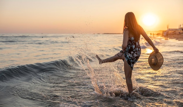 La giovane donna felice in un cappello e in un vestito corto gode del tramonto in mare