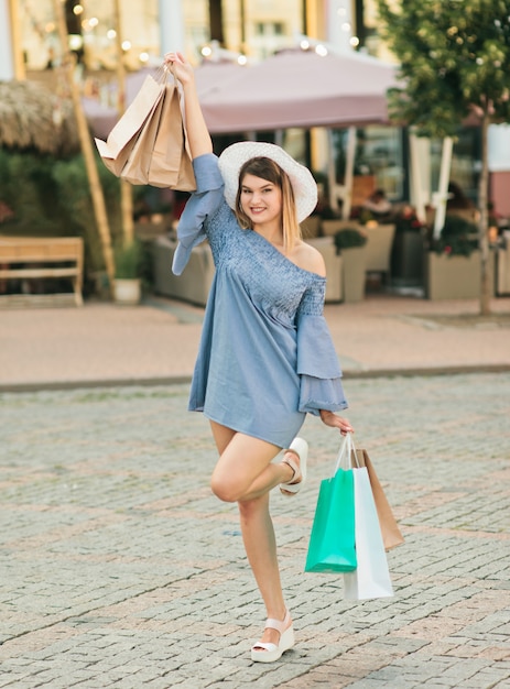La giovane donna felice in un cappello e un vestito tiene i sacchetti della spesa in sue mani e sorride in una città dell'estate