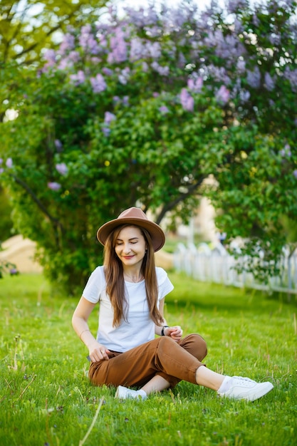 La giovane donna felice in un cappello si siede su un prato verde in un parco. Una ragazza di aspetto europeo con un sorriso sul viso in una luminosa giornata estiva di sole