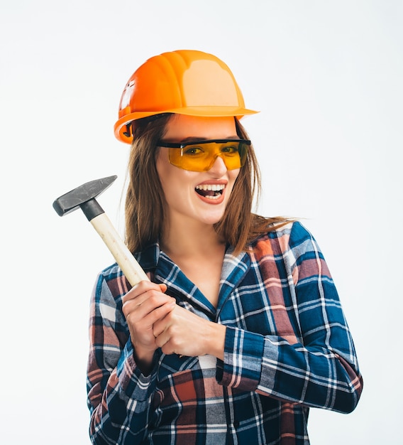 La giovane donna felice indossa un casco di sicurezza arancione, occhiali gialli e camicia a scacchi tenendo il martello a portata di mano.