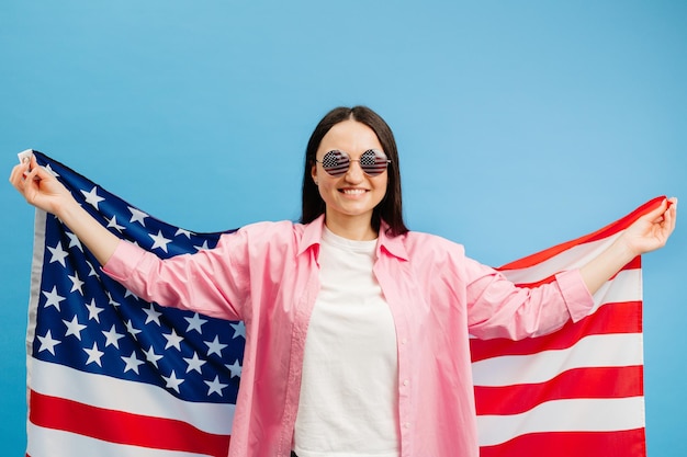 La giovane donna felice sorridente indossa abiti casual tenendo la bandiera americana e guardando la telecamera isolata su sfondo di colore blu