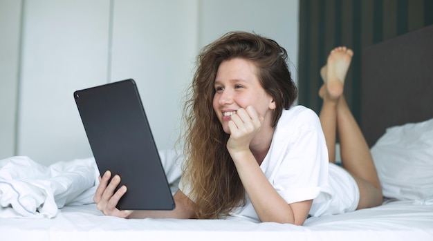 La giovane donna felice sta usando il computer della compressa a casa nel letto nel sorriso della camera da letto si diverte