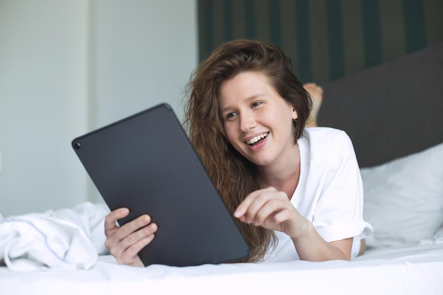 La giovane donna felice sta usando il computer della compressa a casa nel letto nel sorriso della camera da letto si diverte