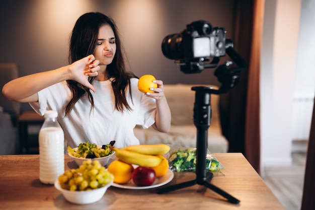 La giovane donna food blogger non ama la frutta fresca e l'insalata nello studio della cucina, mentre riprende il tutorial sulla telecamera per il canale video. L'influencer femminile mostra la preferenza nel cibo, parla di mangiare.