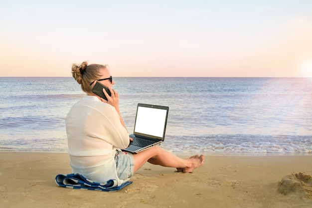 La giovane donna freelance lavora al mare. donna che parla al cellulare, con un computer portatile in mano, sulla spiaggia.