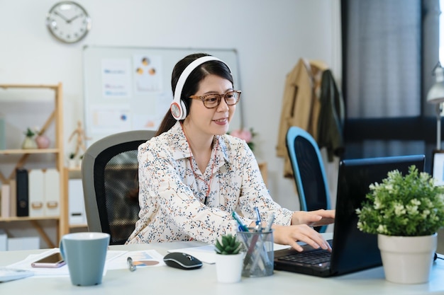 La giovane donna giapponese abbastanza asiatica in occhiali sta lavorando nell'ufficio moderno. rilassare l'impiegata in cuffia ascoltando musica e sorridendo in studio. signora lavoratore digitando sul computer portatile.