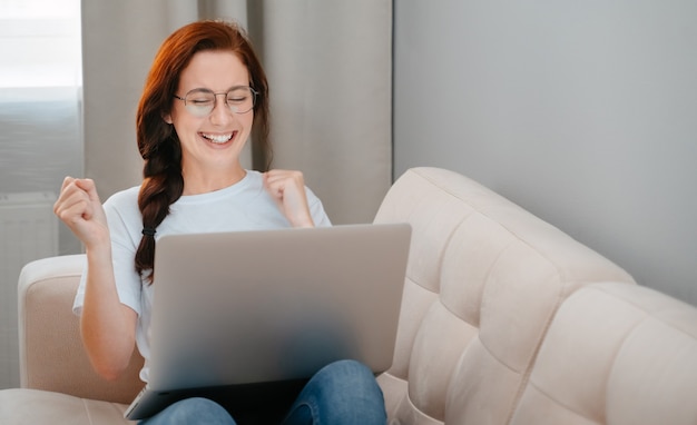 La giovane donna gode del successo guardando lo schermo del laptop mentre è seduta su un comodo divano nel soggiorno...