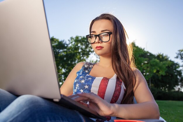 La giovane donna graziosa che si siede sul sacchetto di fagioli usa il computer portatile mentre riposa sull'erba nel parco sul sun