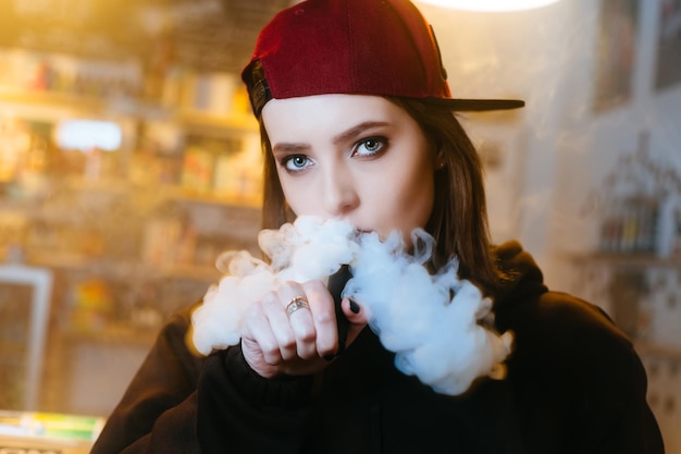 La giovane donna graziosa in berretto rosso fuma una sigaretta elettronica al primo piano di stile hiphop del negozio di vape