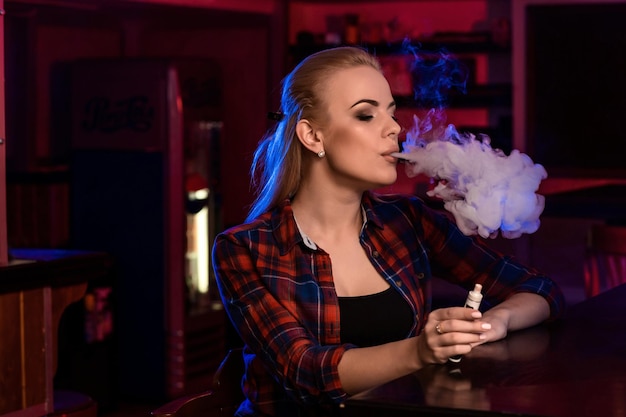 La giovane donna graziosa in una camicia in una gabbia fuma una sigaretta elettronica al bar del vape