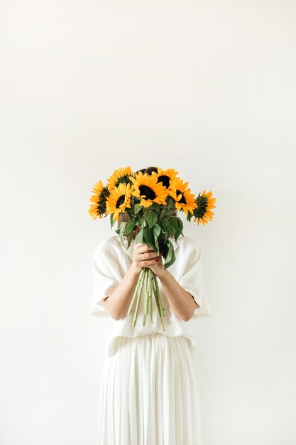 La giovane donna graziosa in vestito bianco tiene il mazzo dei girasoli su bianco