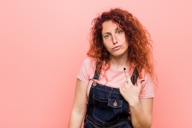 La giovane donna graziosa rossa dello zenzero che indossa un denim dei jeans che indica con il dito voi come se l'invito si avvicini.