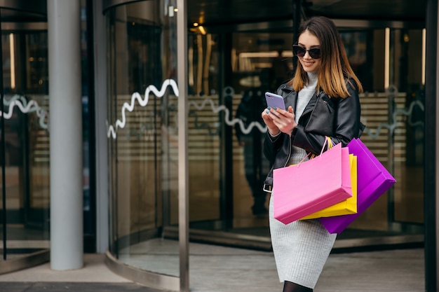 La giovane donna graziosa scarna con il sacchetto della spesa resta vicino al centro commerciale e facendo uso del telefono