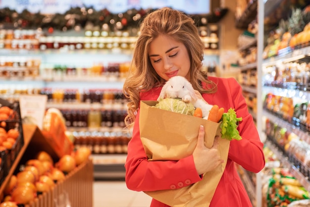 La giovane donna graziosa sta facendo la spesa per comprare le verdure per il suo coniglio bianco da compagnia al supermercato