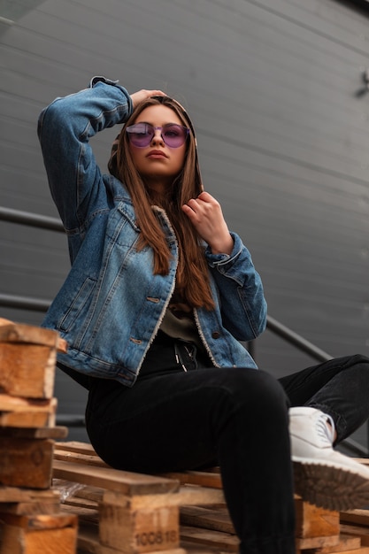 La giovane donna hipster abbastanza americana in vestiti casuali alla moda della gioventù in occhiali viola alla moda posa all'aperto in città. La modella moderna e affascinante della ragazza si siede su pallet di legno in strada.