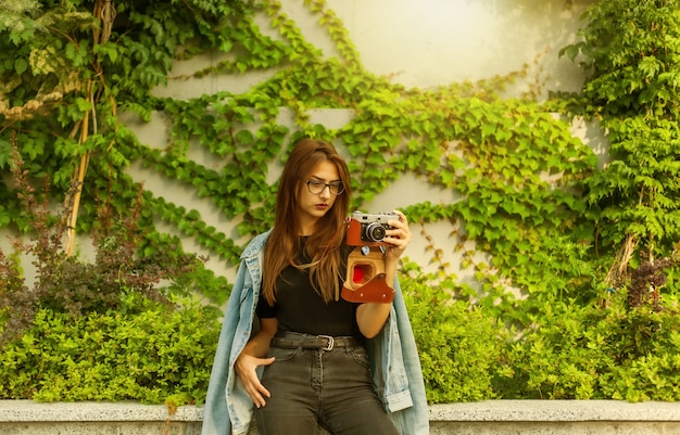 La giovane donna hipster in giacca di jeans e occhiali gode di una telecamera retrò all'aperto