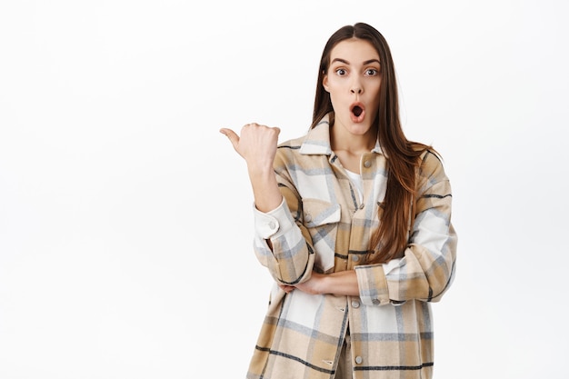 La giovane donna impressionata indica a sinistra il logo dello spazio della copia, guarda con la faccia eccitata davanti, in piedi in abito elegante contro il muro bianco