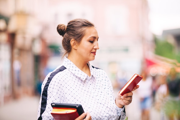La giovane donna in camicia bianca tiene il tablet e il taccuino è in piedi in città e guarda lo smartphone