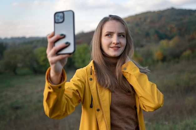 La giovane donna in giacca gialla gode della natura autunnale Ragazza sorridente che prende selfie sullo smartphone sullo sfondo del paesaggio di montagna