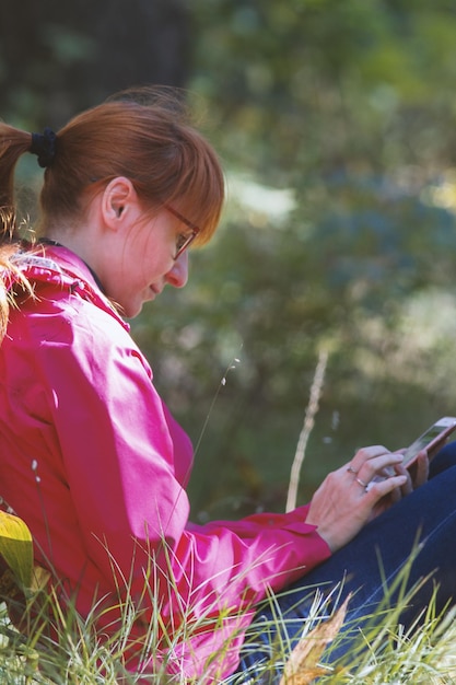 La giovane donna in giacca rosa utilizza lo smartphone nella foresta autunnale - verticale, teleobiettivo