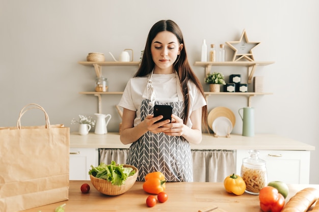 La giovane donna in grembiule usa lo smartphone nella cucina moderna che prepara insalata legge la ricetta