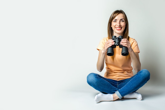 La giovane donna in maglietta e jeans sorride mentre si siede sul pavimento e tiene il binocolo su una priorità bassa chiara