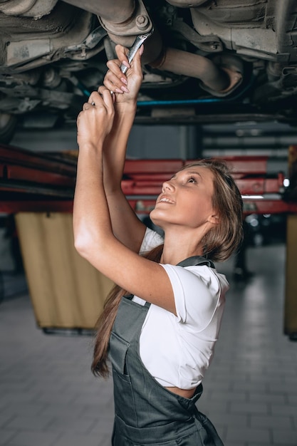 La giovane donna in tuta nera e maglietta bianca svita il dado con una chiave inglese sotto l'auto e sorride. il meccanico donna lavora sotto la parte inferiore di un'auto