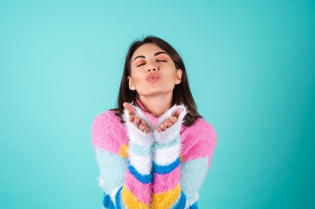 La giovane donna in un maglione multicolore luminoso sull'azzurro invia i baci dell'aria alla macchina fotografica