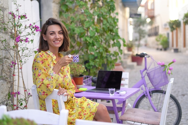 La giovane donna in un vestito in un caffè luminoso della via con un computer portatile beve caffè turco, lavora a distanza sul suo proprio programma