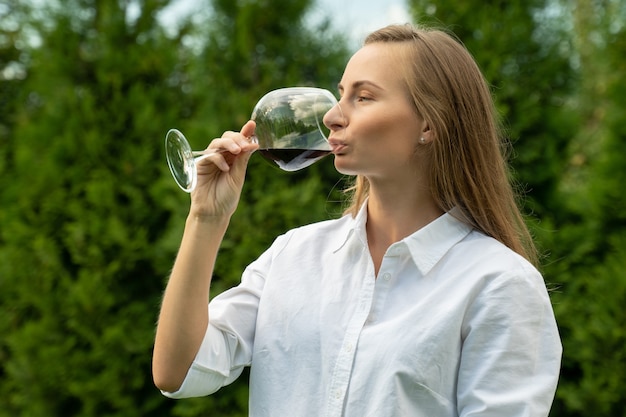 La giovane donna in una camicia bianca annusa e assaggia il vino rosso su un fondo verde