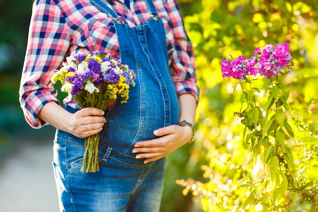 La giovane donna incinta in tuta del denim tiene i fiori