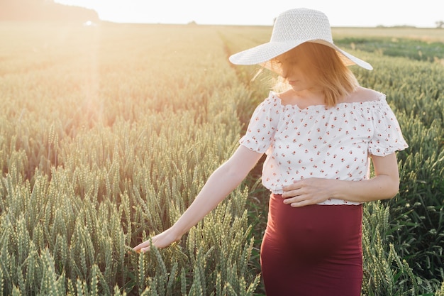 La giovane donna incinta mentre cammina nei campi accarezza le cime dei cereali coltivati con la mano. Cura della gravidanza. Bellezza e salute. Felicità e serenità. Uno stile di vita sano. Prodotti per donne in gravidanza.