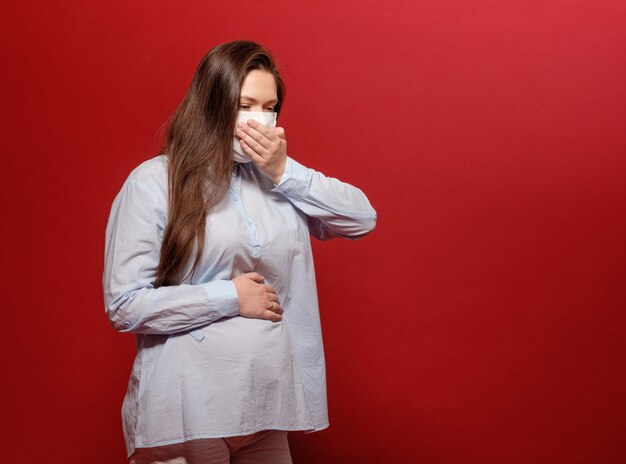 La giovane donna incinta sulla parete rossa in mascherina medica protettiva tiene il suo stomaco e starnutisce