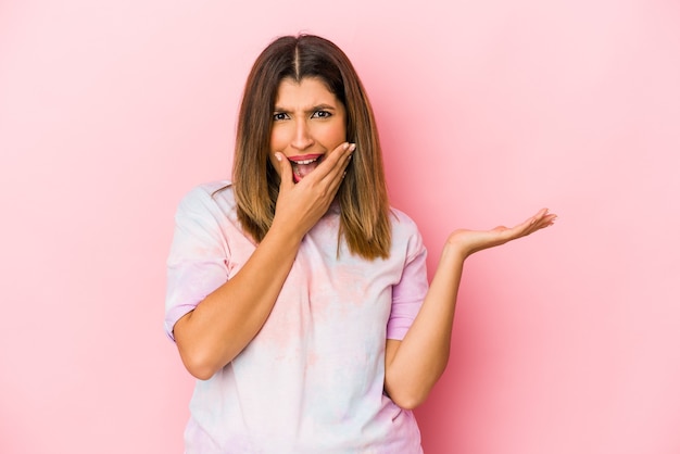 La giovane donna indiana isolata su fondo rosa tiene lo spazio della copia su una palma, tiene la mano sulla guancia. Stupito e deliziato.