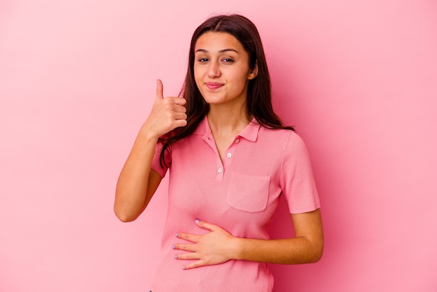 La giovane donna indiana isolata sulla parete rosa tocca la pancia, sorride delicatamente, mangia e concetto di soddisfazione.