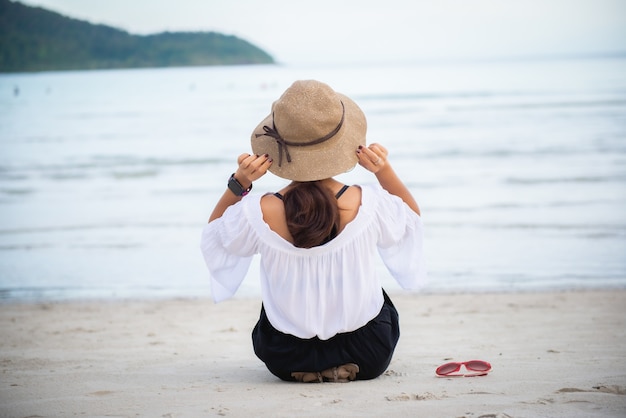 La giovane donna indossa un cappello seduto sulla spiaggia di fronte al mare e ha gli occhiali posizionati sul lato.