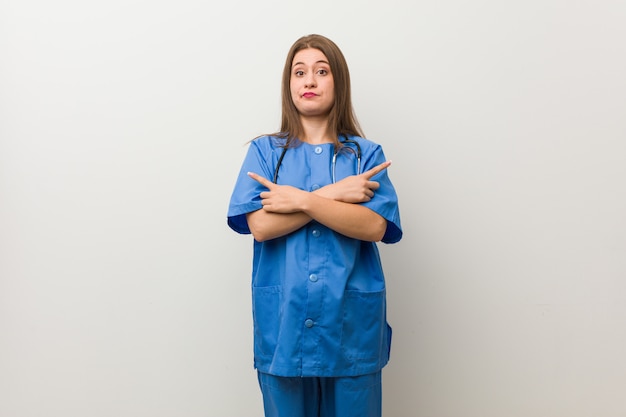 La giovane donna infermiera contro un muro bianco punta lateralmente, sta cercando di scegliere tra due opzioni.