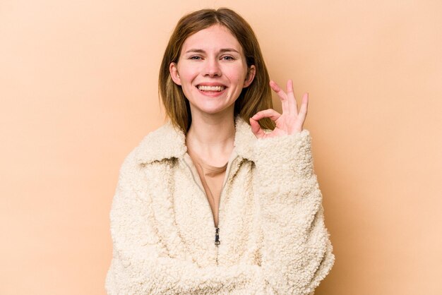 La giovane donna inglese isolata su fondo beige strizza l'occhio e tiene un gesto giusto con la mano.