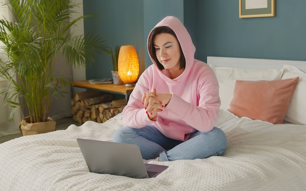 La giovane donna insolita si siede su un letto a casa accogliente e fare una videoconferenza. Ragazza attraente in chat, sorridente, ridere e parlare. Comunicazione online sul laptop.