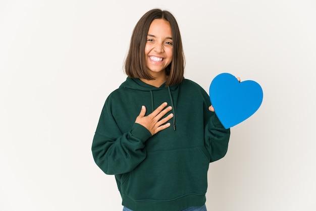 La giovane donna ispanica che tiene una figura del cuore ride ad alta voce mantenendo la mano sul petto.